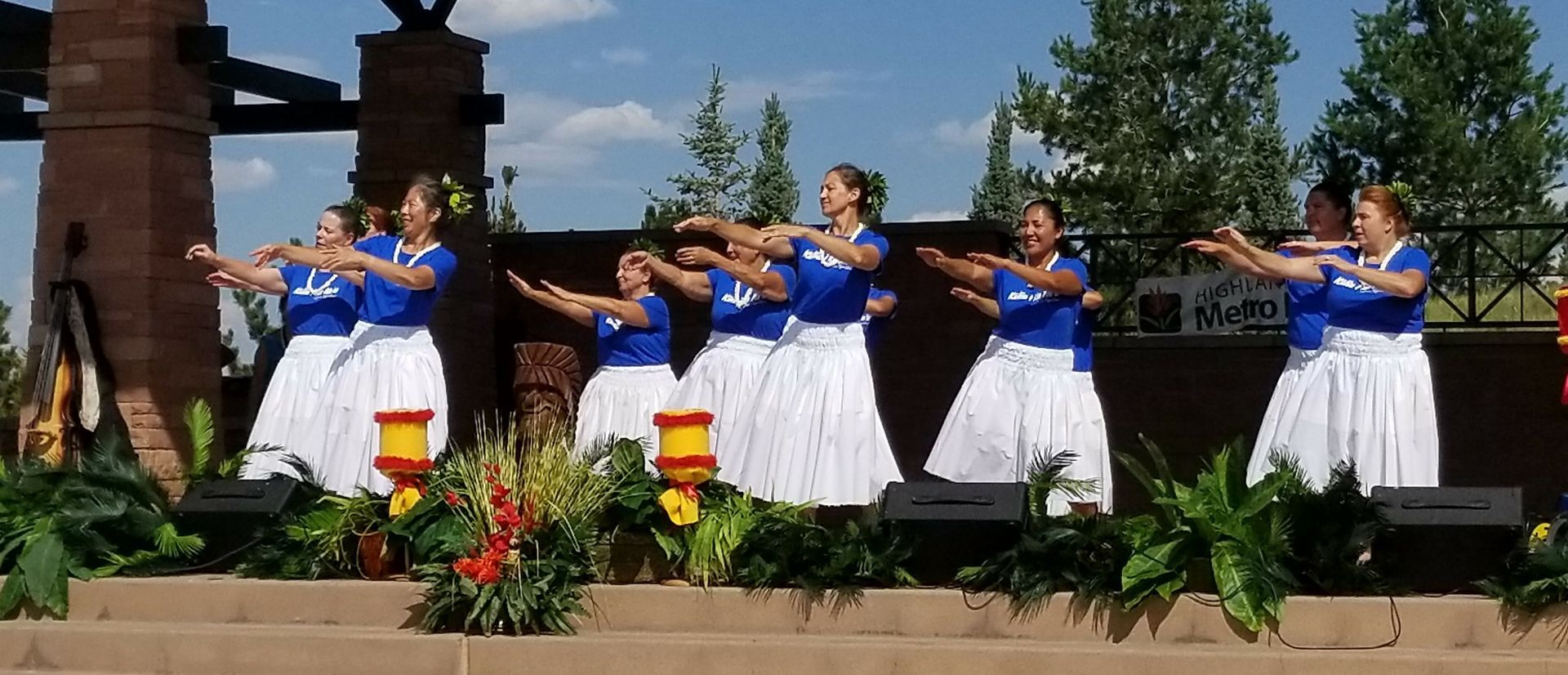 Dancers in blue tops and white skirts perform with arms extended on an outdoor stage decorated with tropical plants.