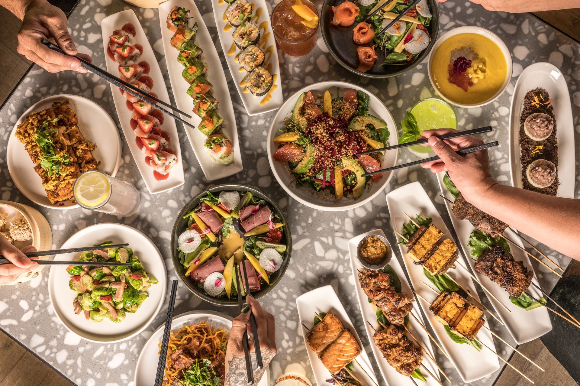 People dining at a table with various Asian dishes, including sushi, salads, and skewers.