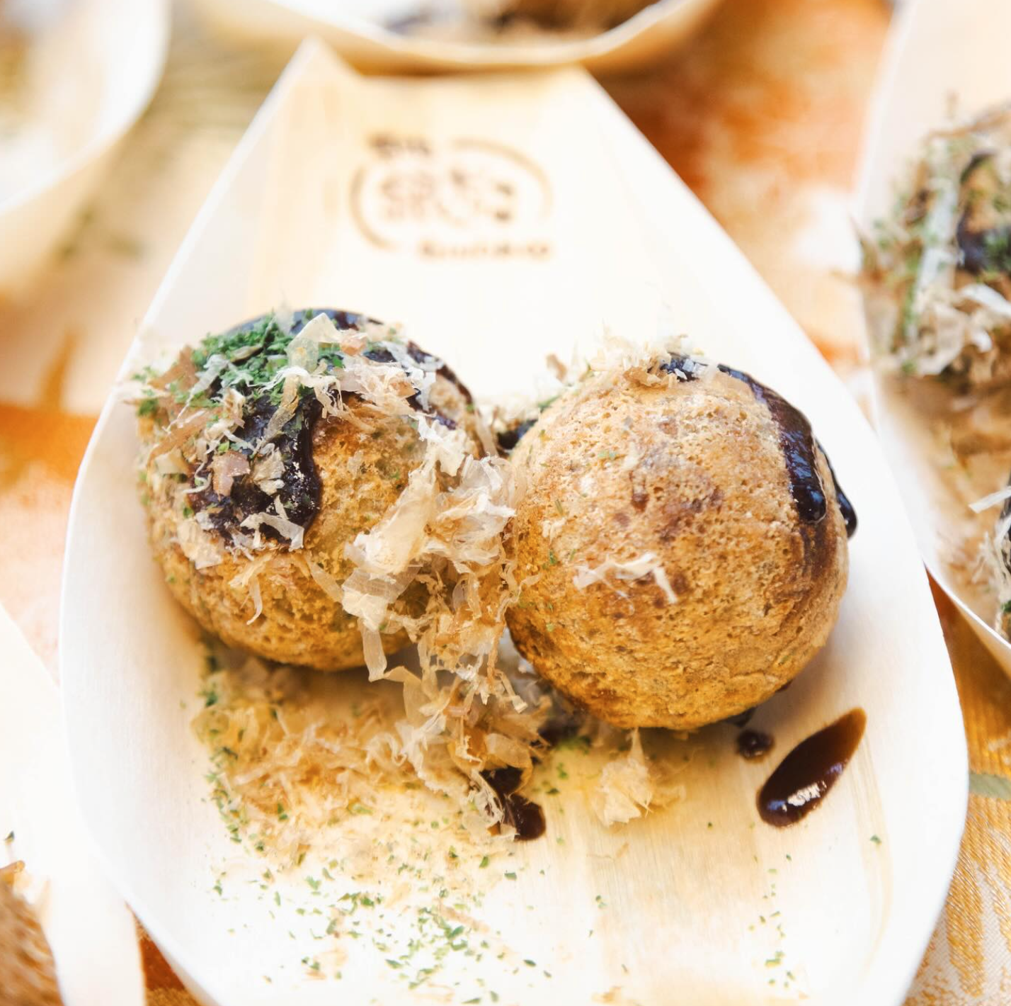 Two takoyaki balls on a leaf-shaped plate, topped with sauce, flakes, and herbs.