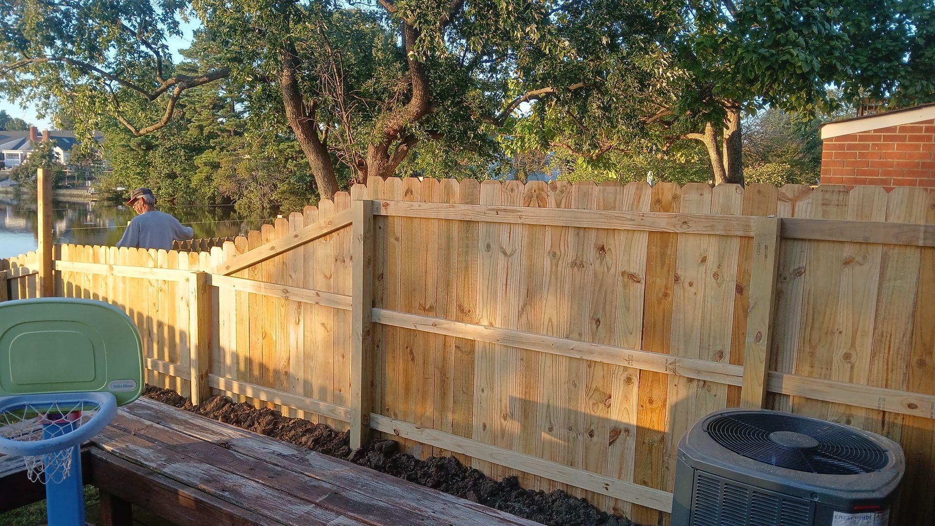 Wooden fence in sunlight with person in the background, a green chair, and a trash can on a deck.