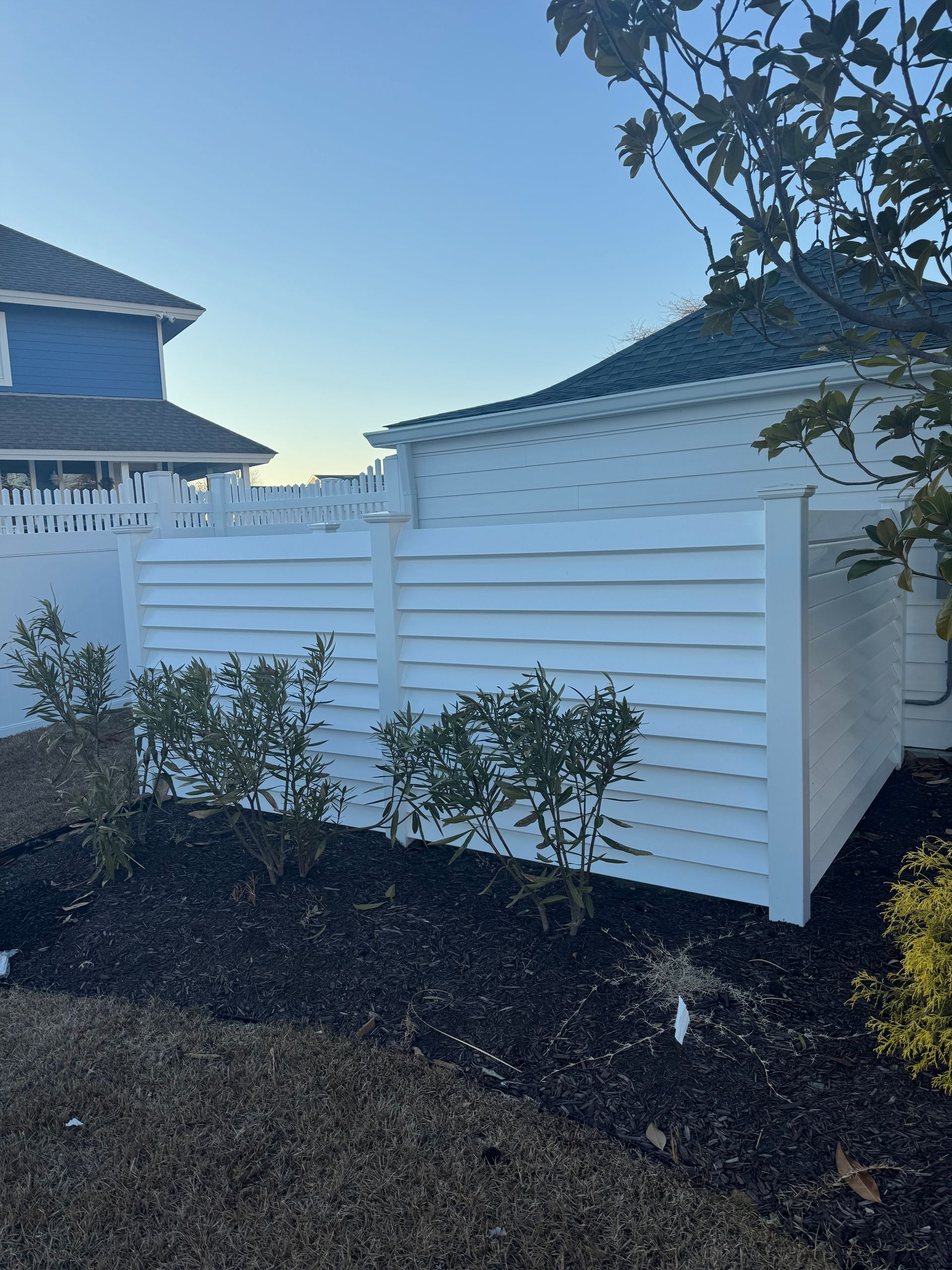 White slatted fence near small bushes and dark mulch, with part of a blue-roofed building visible.