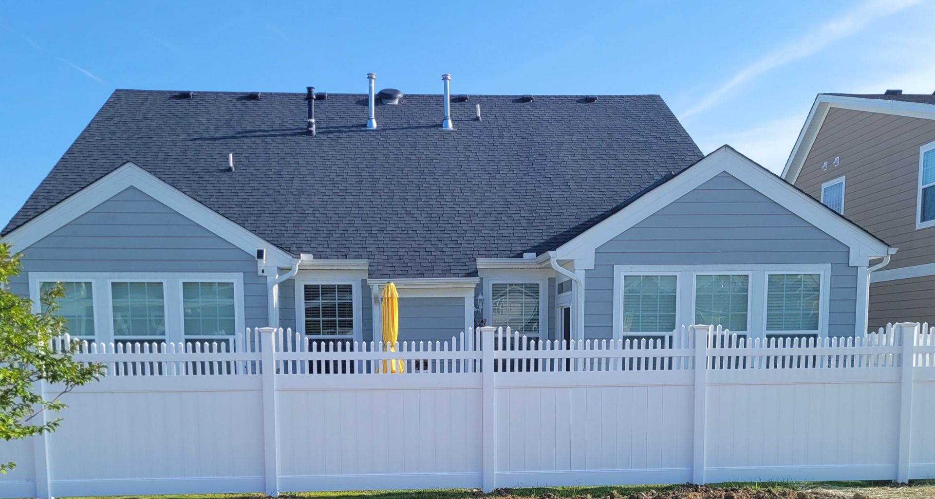 Rear view of a light blue house with a dark gray roof behind a white picket fence, under a blue sky.