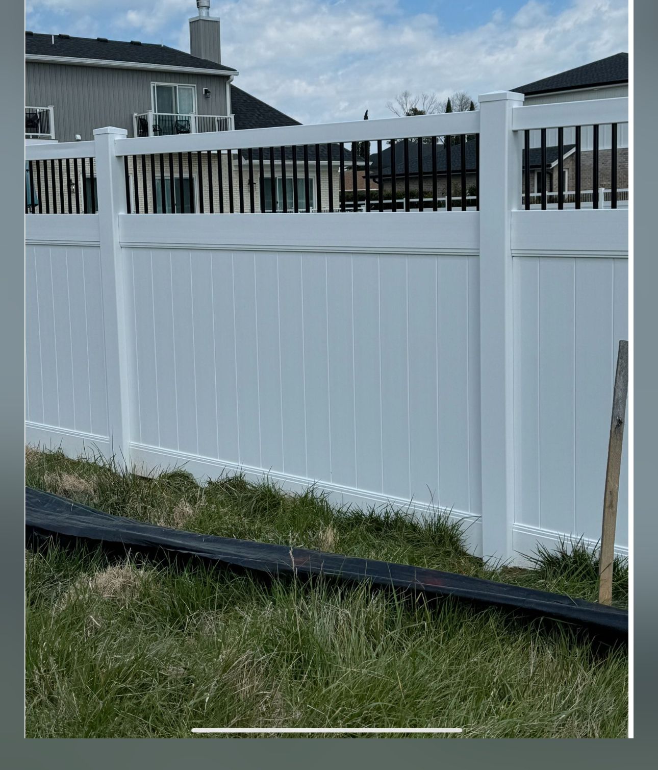 White vinyl fence with black metal railing on top, in a yard with grass and blue sky.