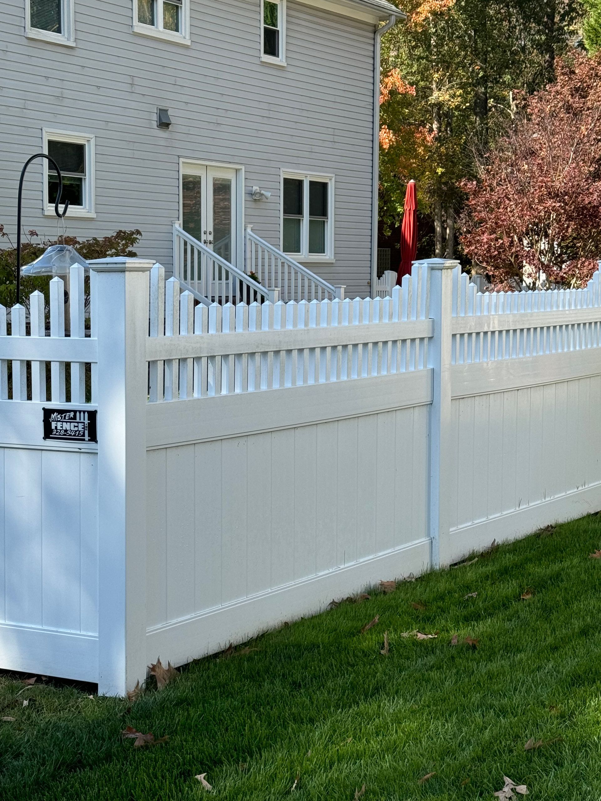 White picket fence in front of a light gray house with green grass and trees in the background.