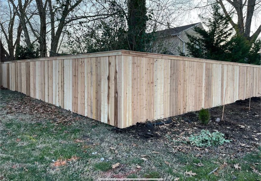 Wooden fence corner with vertical boards, framing a yard with grass and trees.