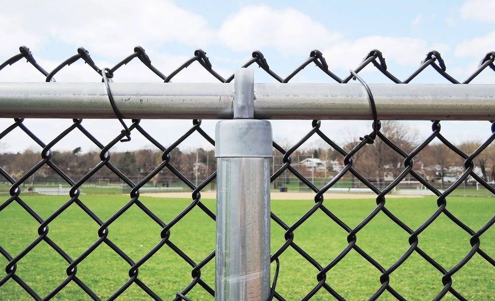Chain link fence, grey post, black mesh, green field and trees in the background under a blue sky.