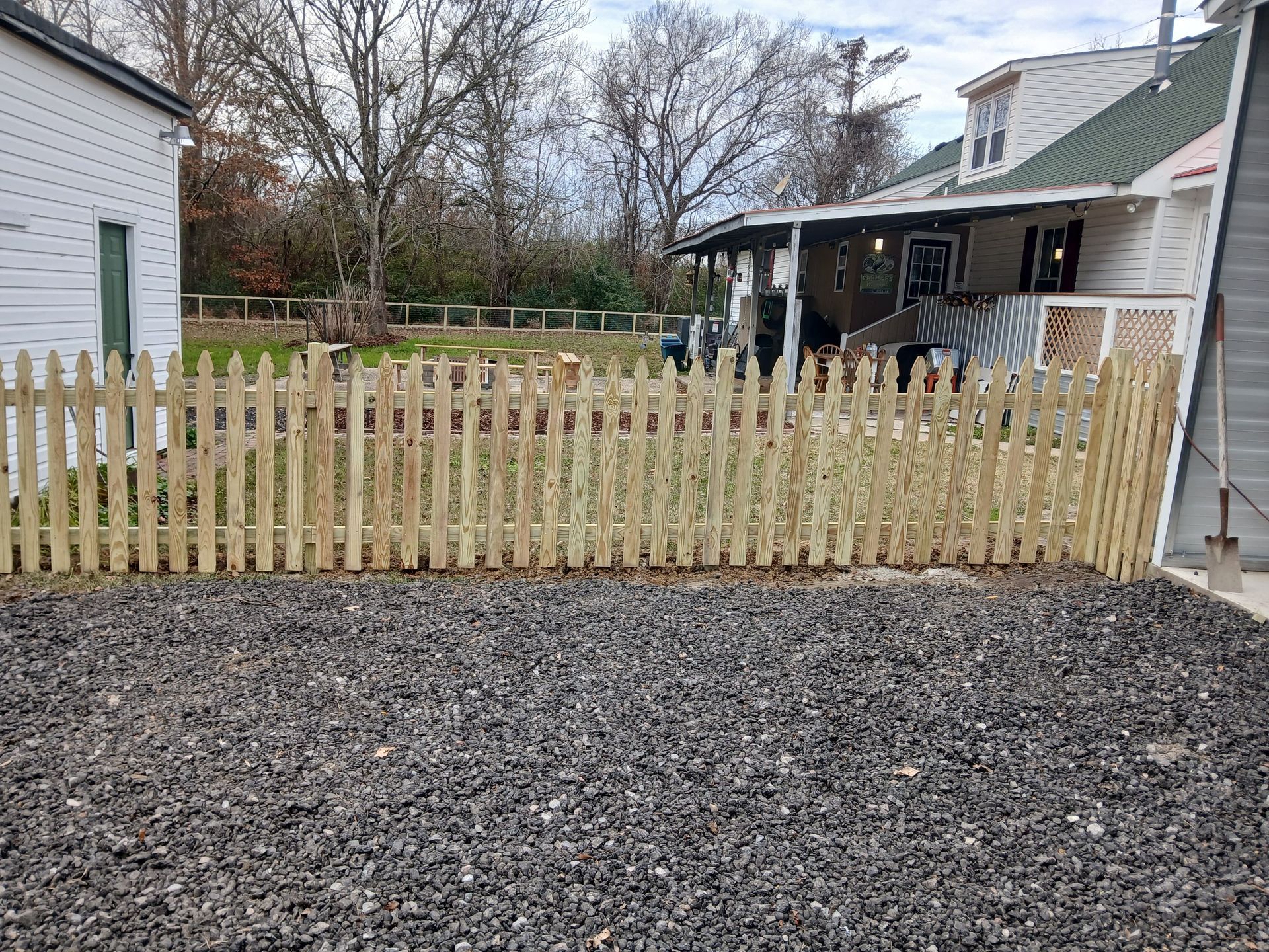 A wooden picket fence encloses a gravel yard in front of a house, with another house visible in the background.