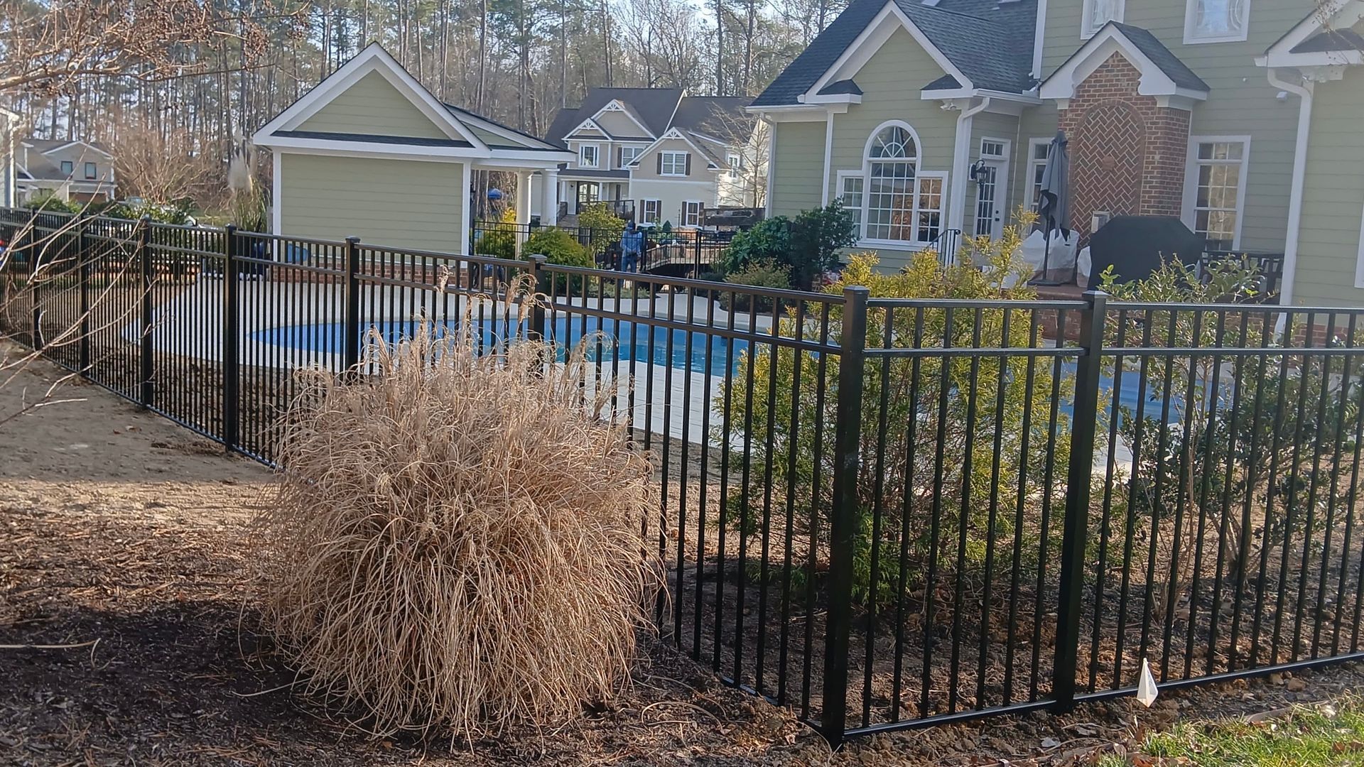 Black fence surrounds a swimming pool behind a green house.  Brown foliage in the foreground.