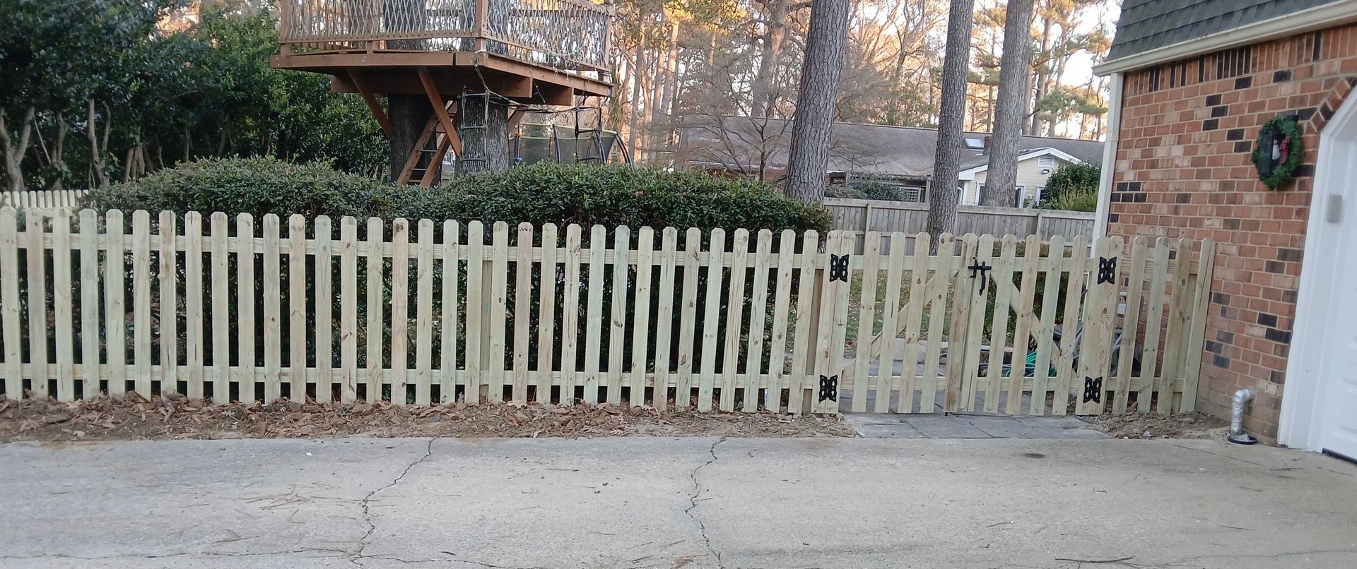 Light wooden picket fence in front of greenery and a brick building, a treehouse visible in the background.