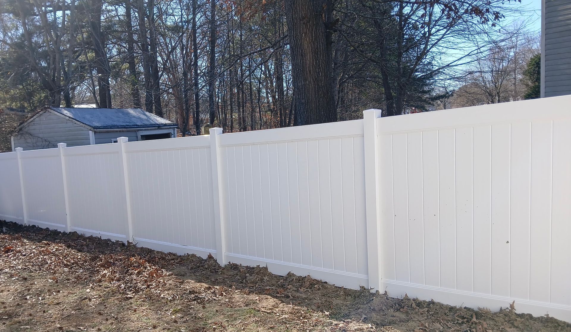 White vinyl fence in front of a yard with a small building in the background. Brown leaves are in the yard.