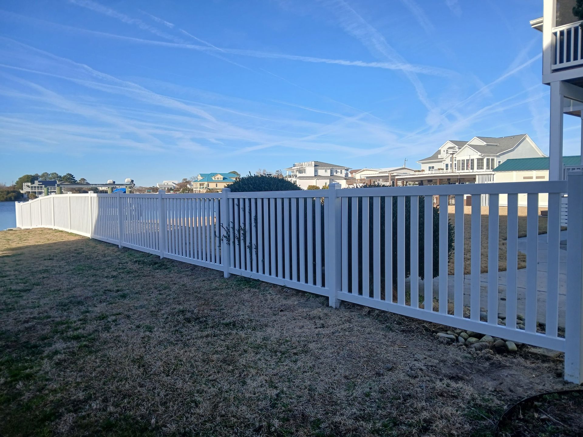 White picket fence on grassy lawn, waterfront houses in the background under a blue sky.