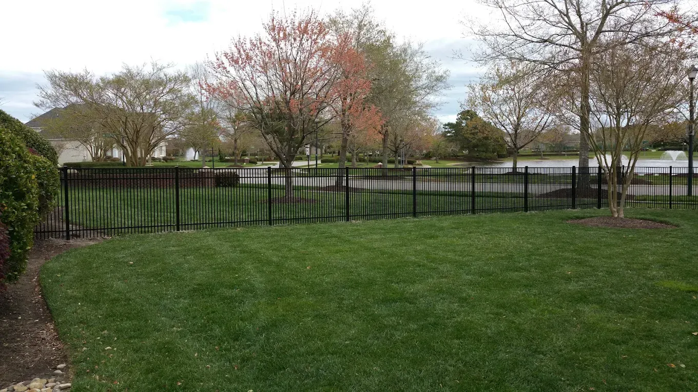 Lush green lawn with black metal fence bordering a body of water, trees in background under a cloudy sky.