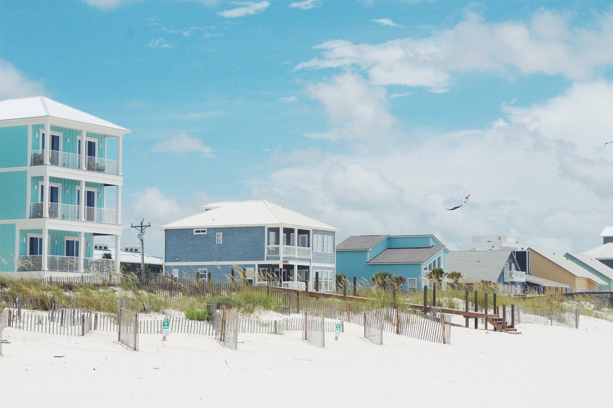Multi-story colorful beach houses sit behind sand dunes and white sand under a blue sky with soft clouds.