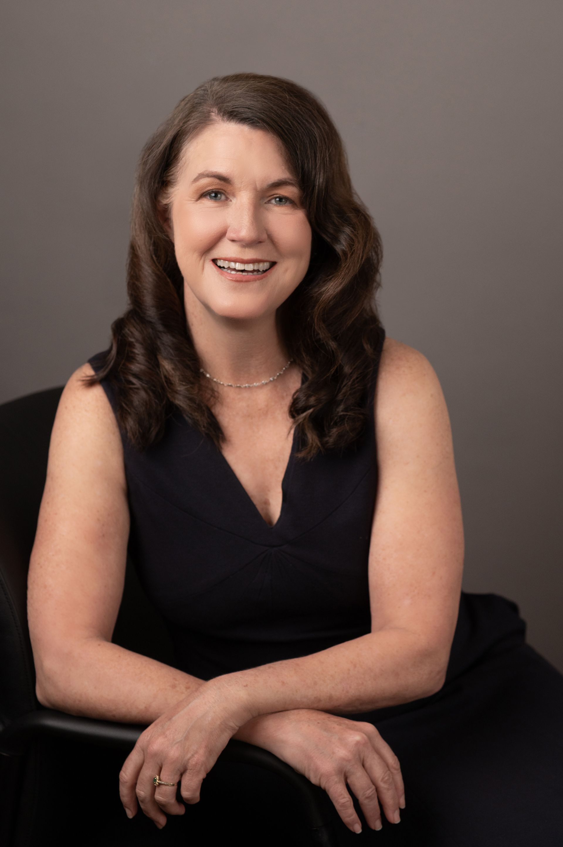A woman in a black dress is sitting in a chair with her legs crossed and smiling.