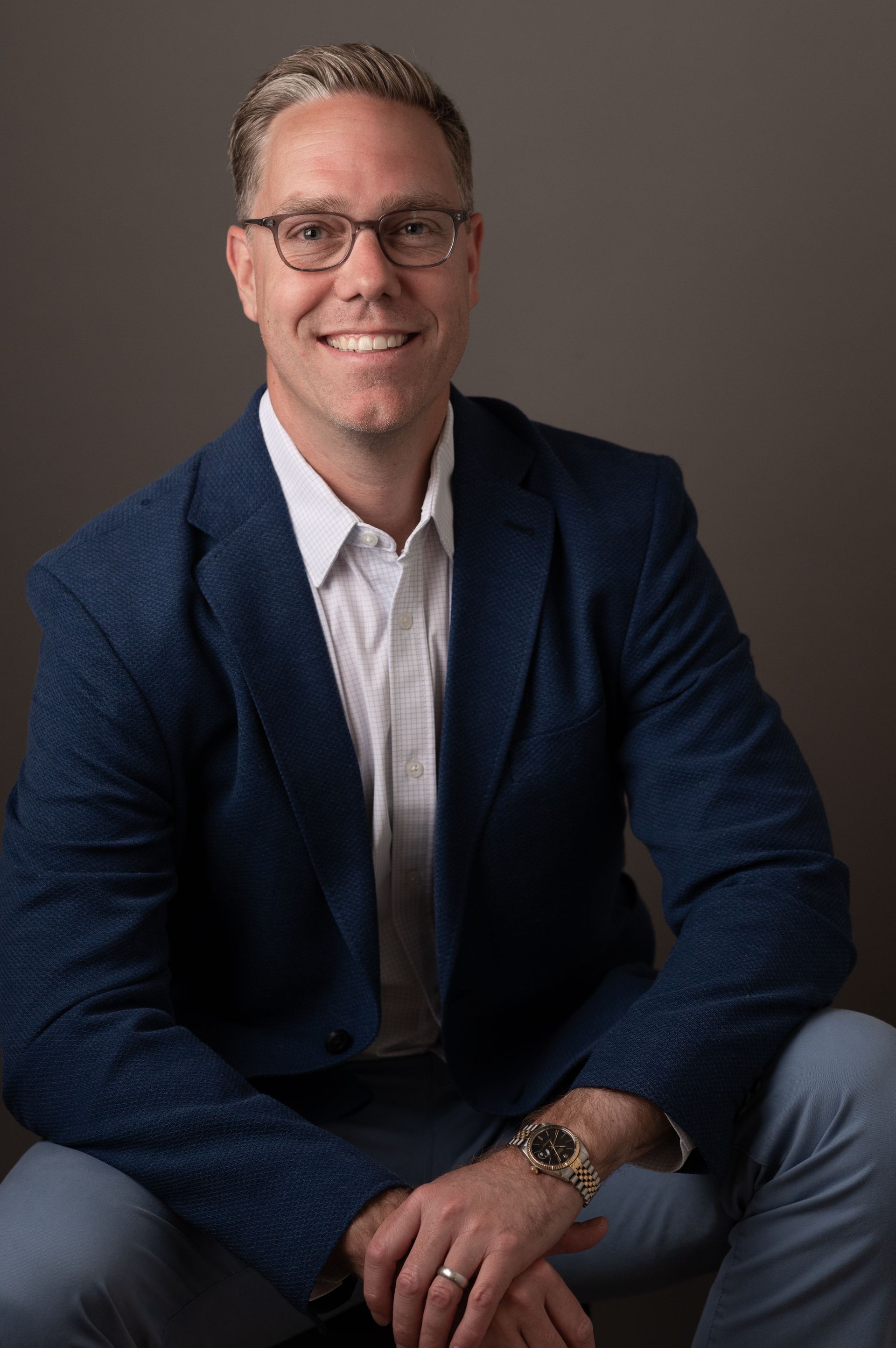 A man wearing glasses and a blue jacket is sitting down and smiling.