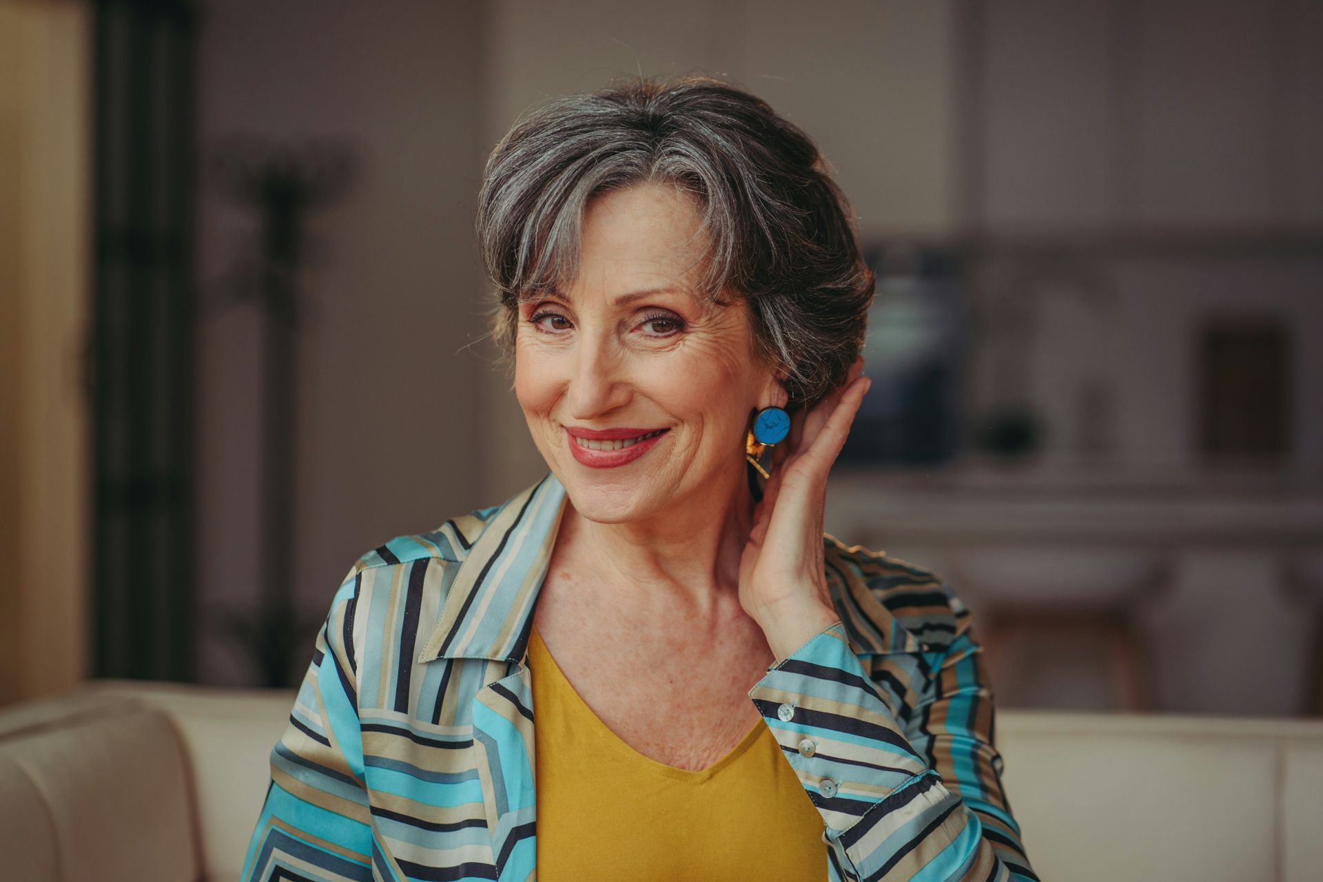 An elderly woman is sitting on a couch and smiling while touching her ear.