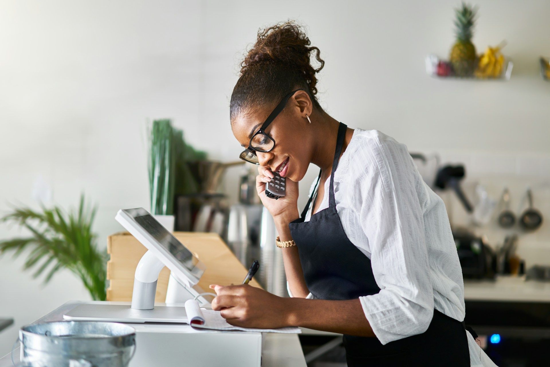 A woman is talking on a cell phone while taking notes in a restaurant.