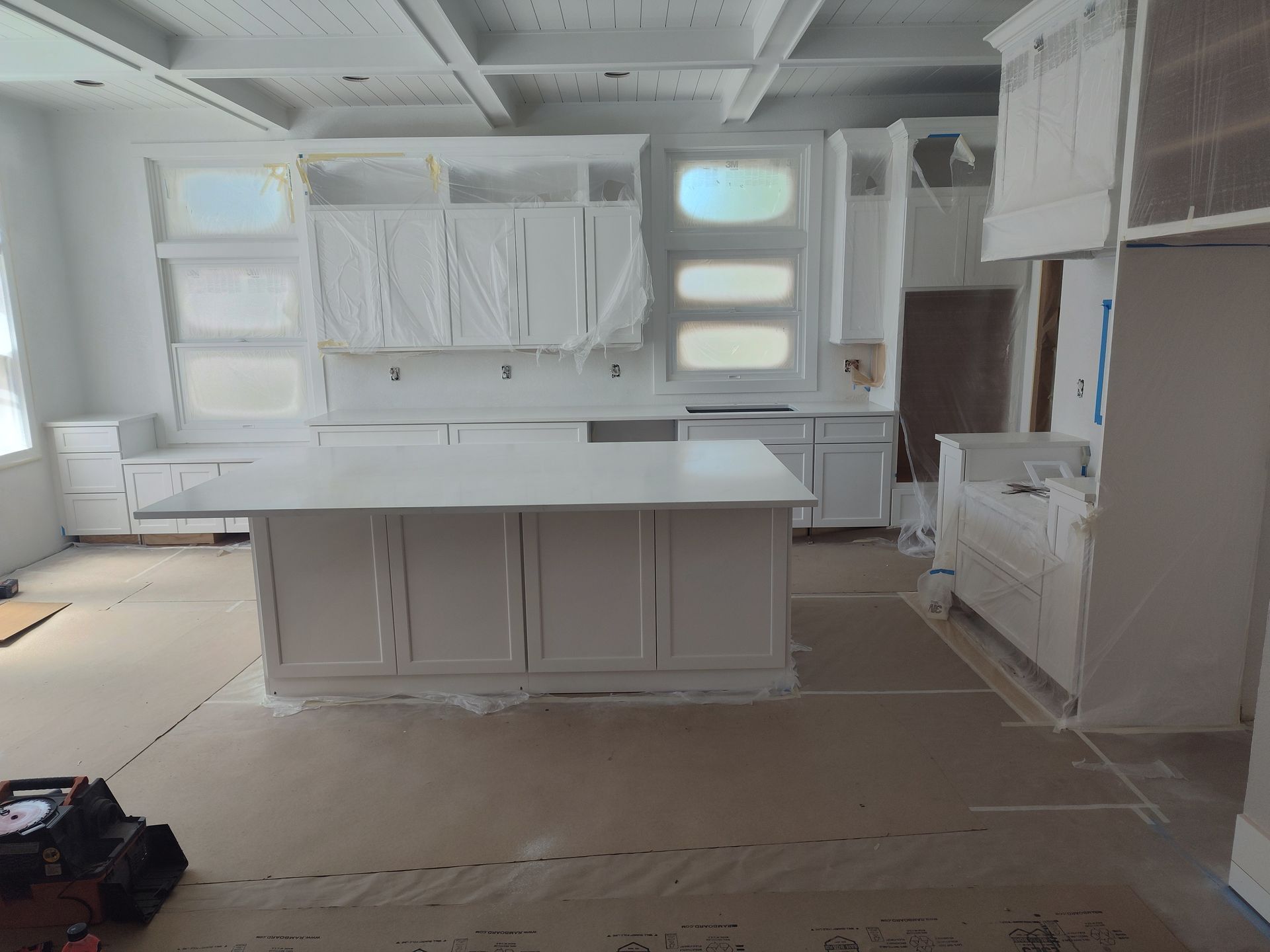 White kitchen under construction, covered in plastic. Island in the center, cabinets on walls.
