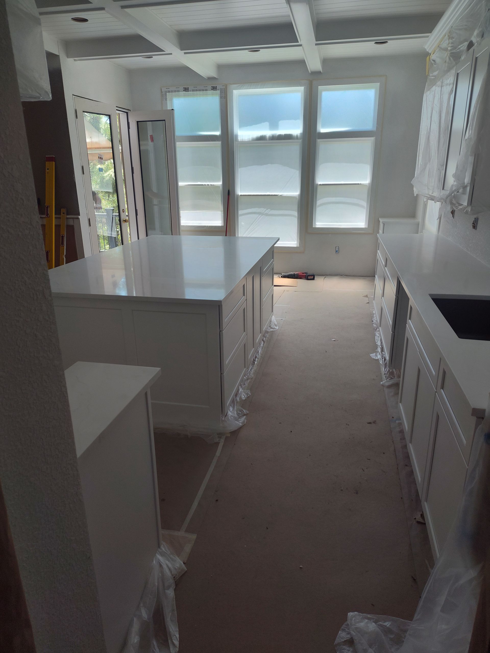 A white kitchen under construction. Island, counters, and cabinets are present. Windows above.