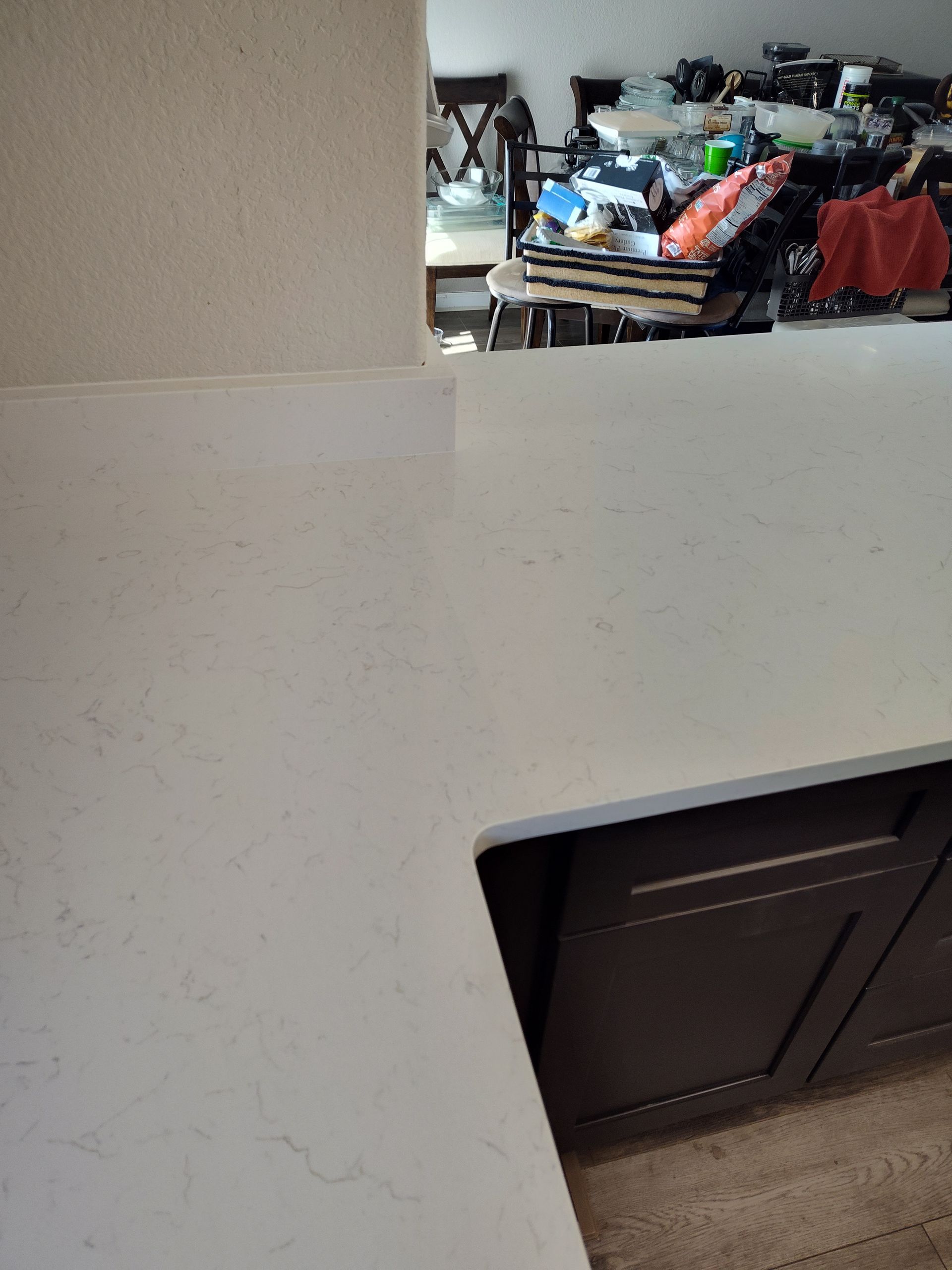White countertop with dark brown cabinets. In background, a cluttered room is partially visible.