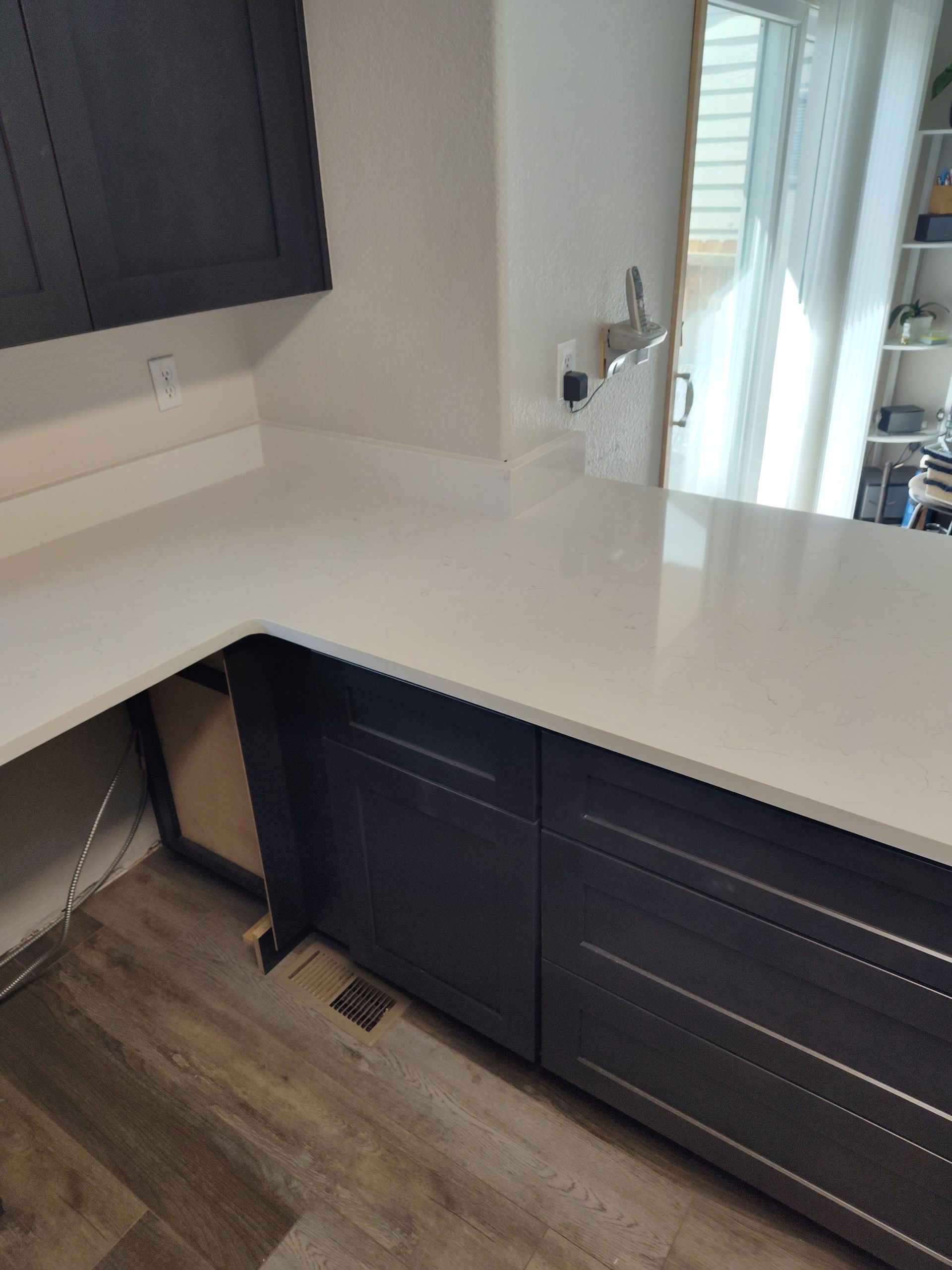 Dark cabinets with white countertop in a kitchen. Gray flooring and window in background.