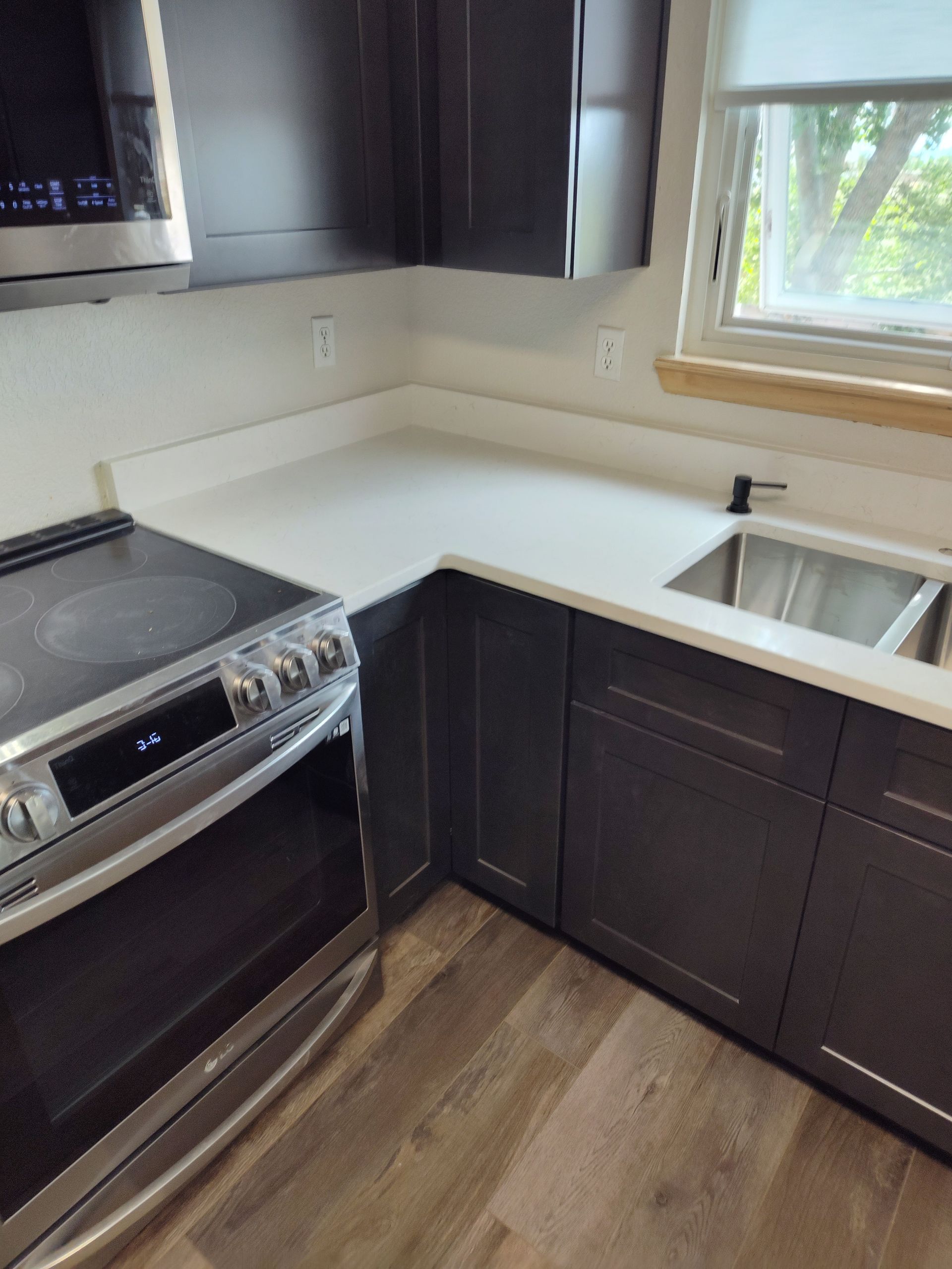 Modern kitchen corner with dark cabinets, white countertops, stainless steel appliances, and a window.
