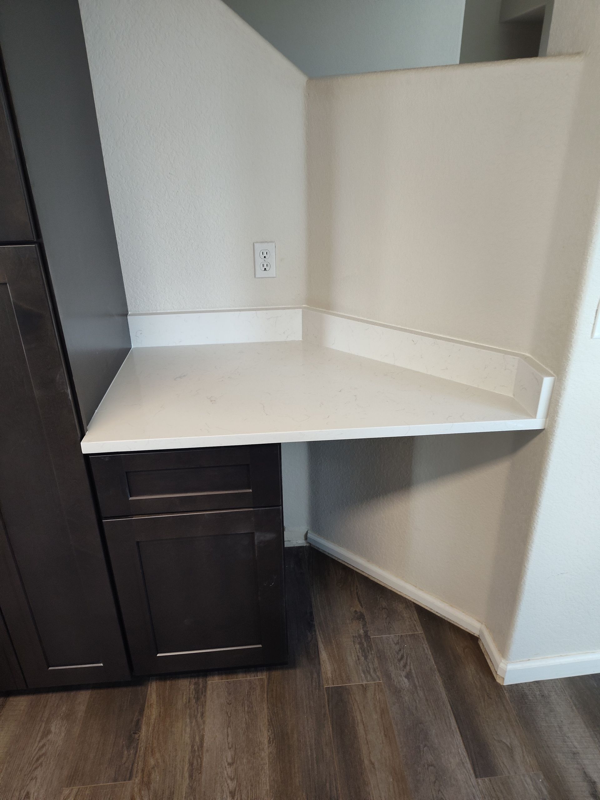 Dark brown kitchen cabinets with white countertop and corner desk area, set on gray-brown wood-look flooring.
