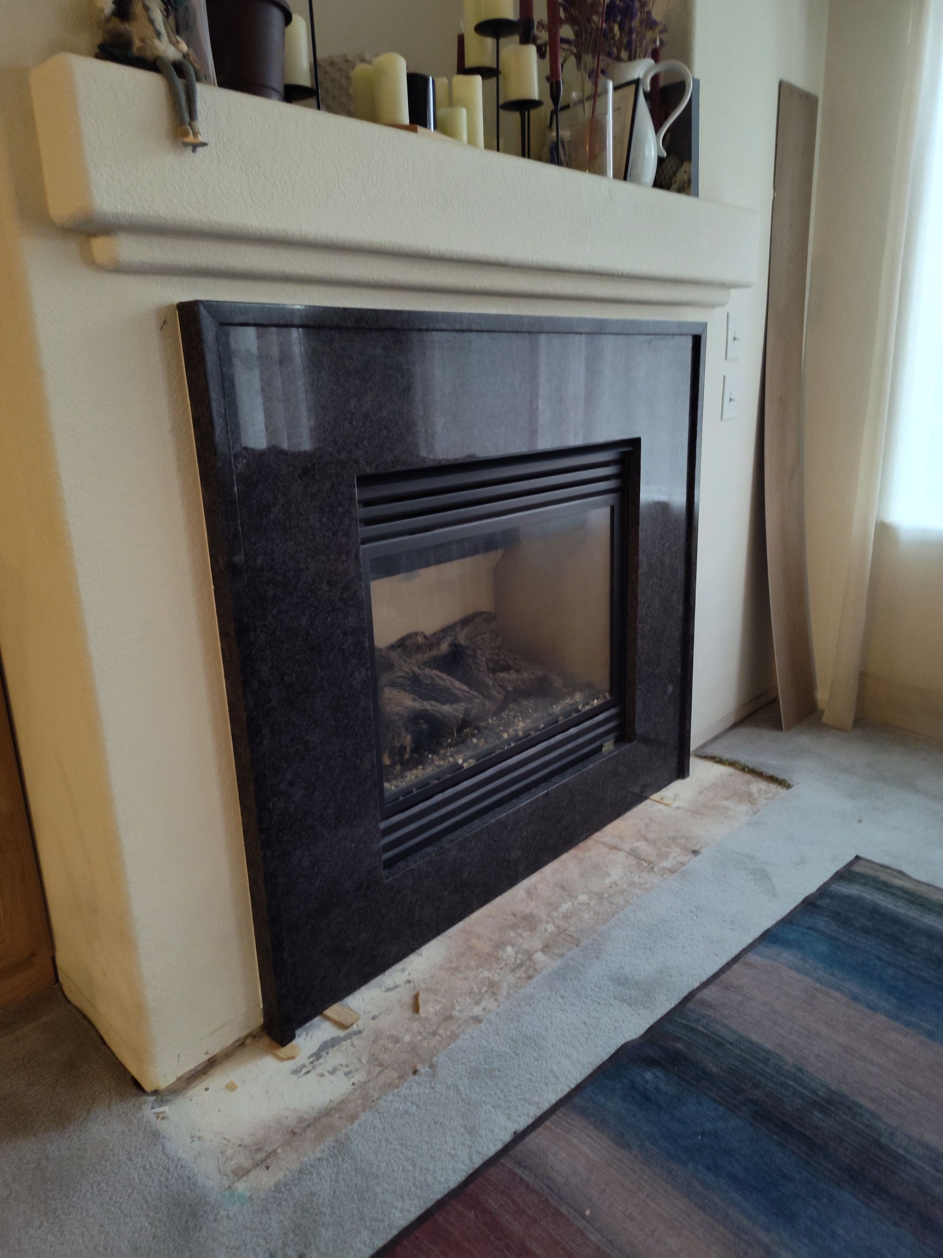 Fireplace with black marble surround, beige mantel, candles, rug, and a partial view of a window.