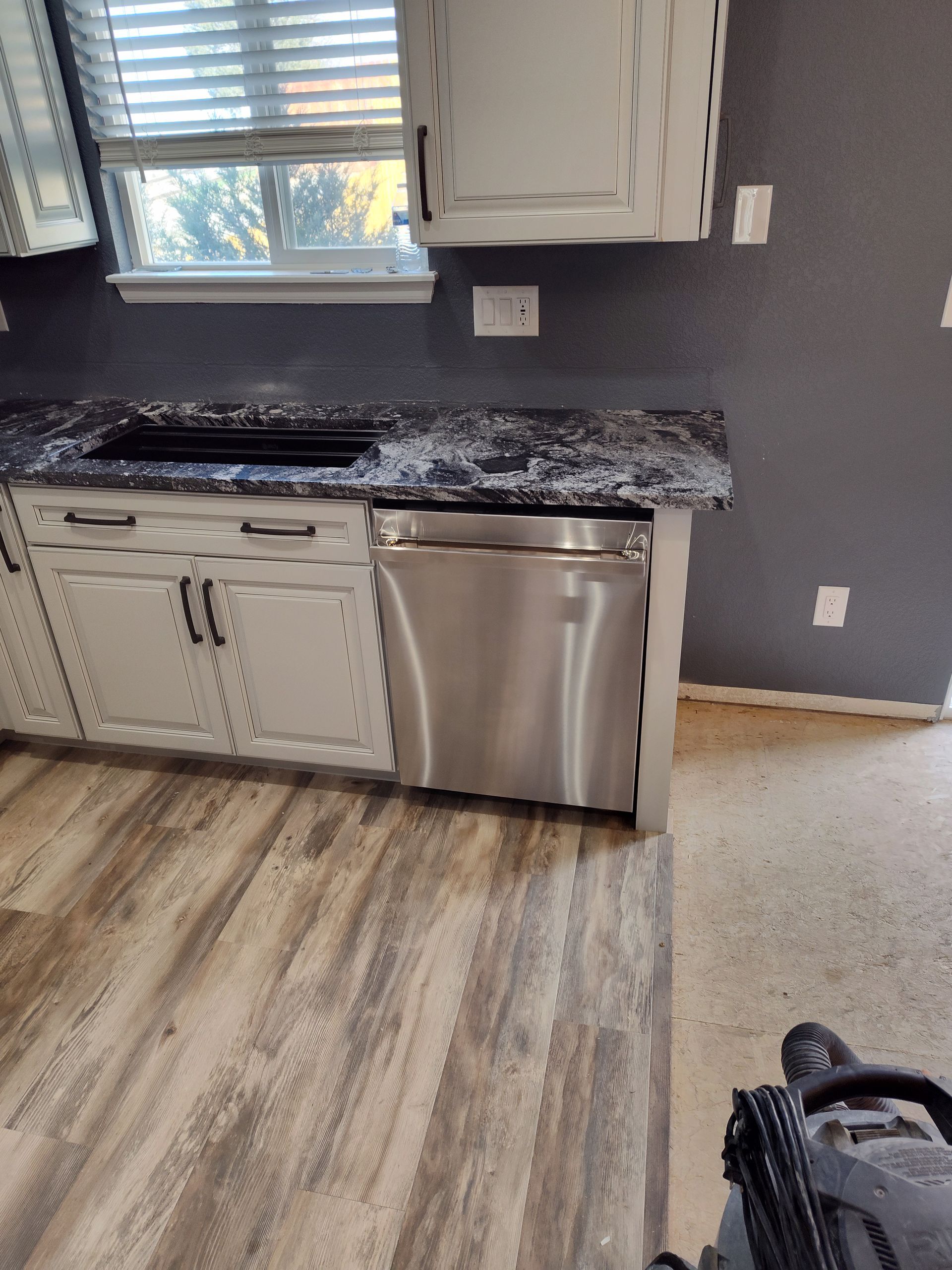 Kitchen with stainless steel dishwasher, gray cabinets, black countertop, and wood-look flooring.