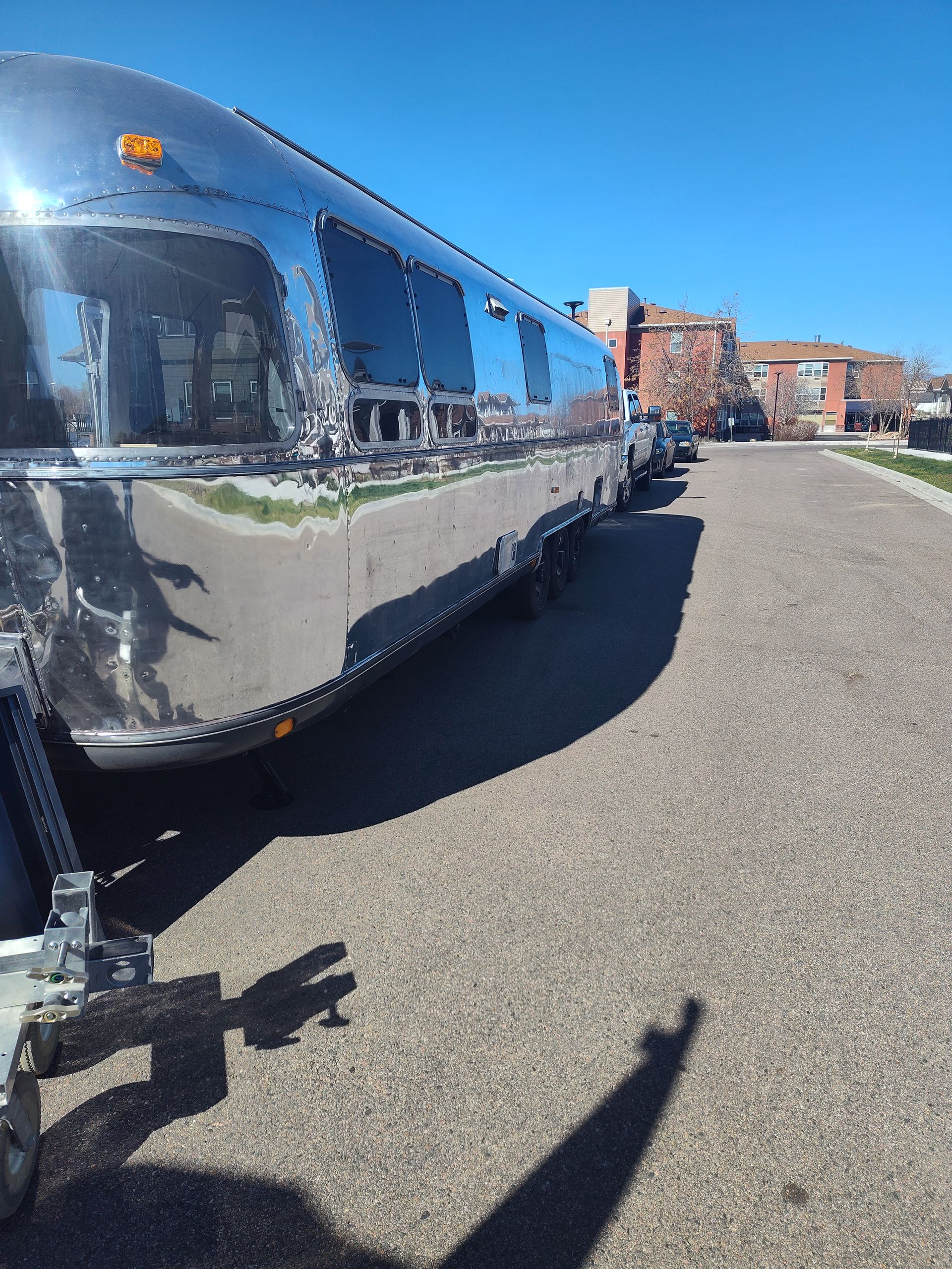 Shiny silver Airstream trailer parked on a paved street, casting a long shadow on a sunny day.
