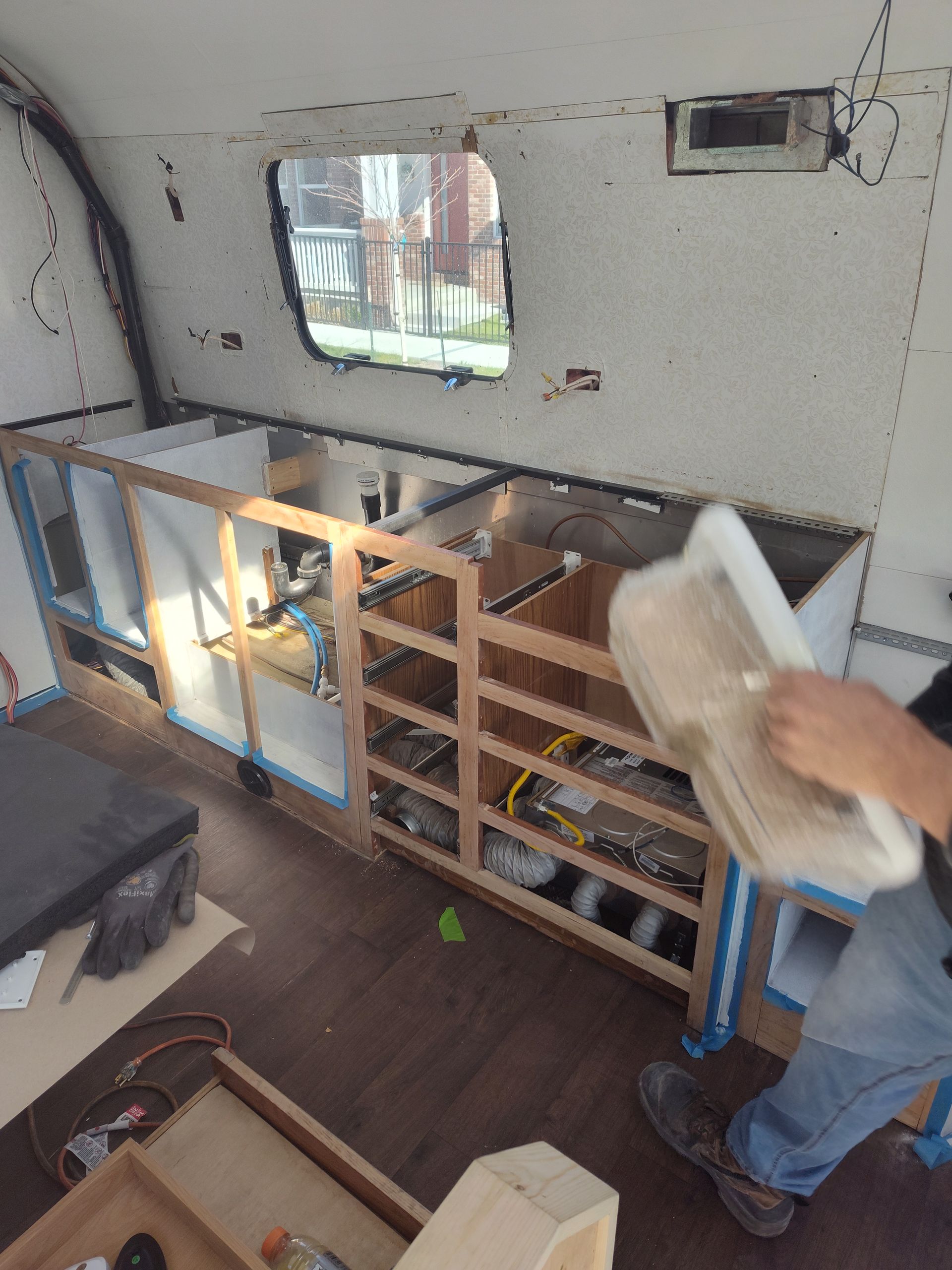 Interior of a renovated camper. A person plasters a wall near a built-in cabinet and window.
