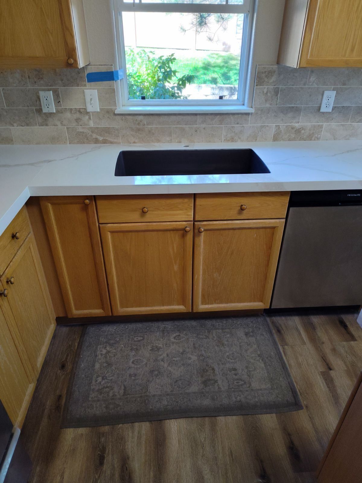 Kitchen with light cabinets, white countertop, black sink, stainless steel dishwasher, and a window.
