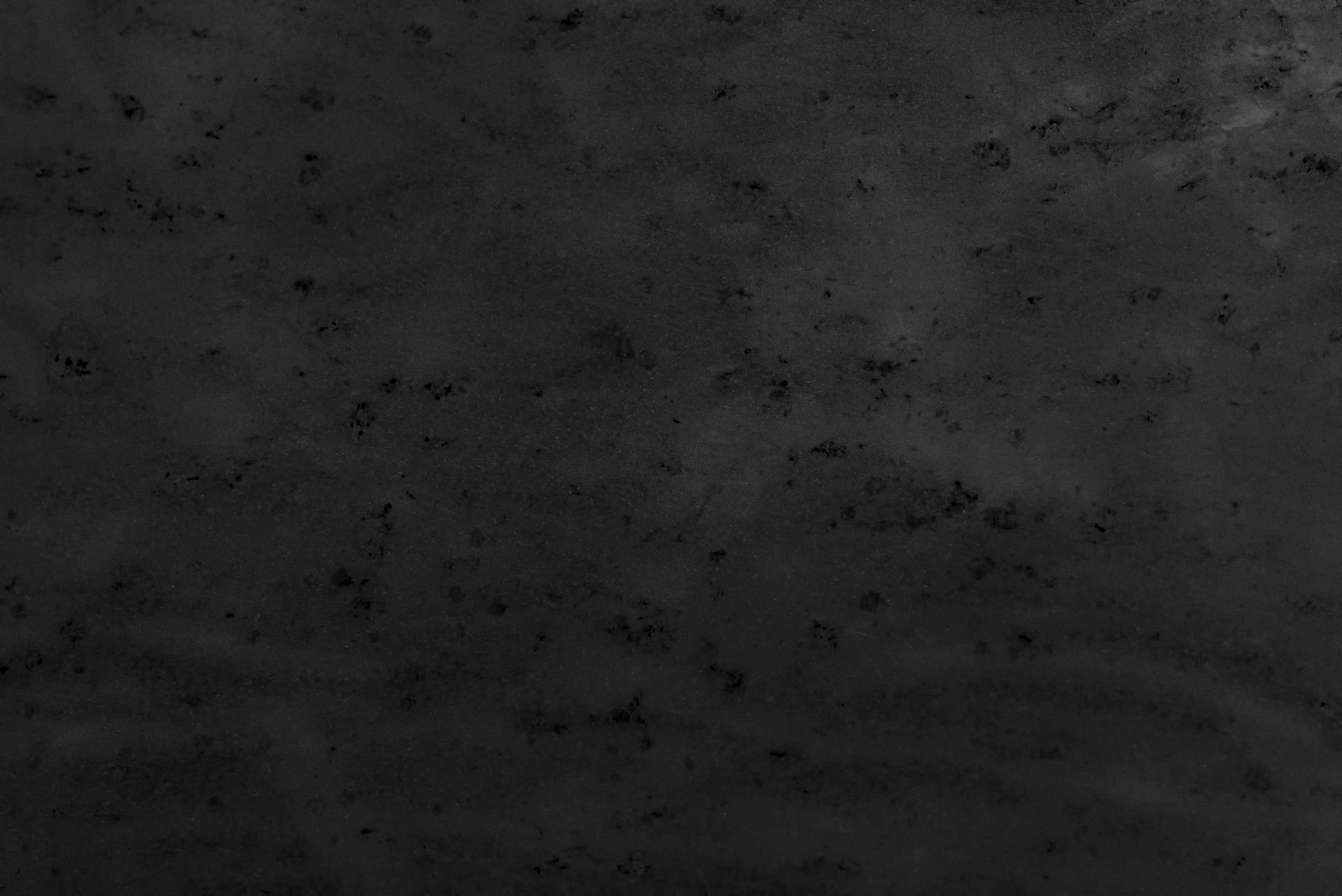 Dark, textured surface with mottled appearance, showing varying shades of gray and subtle patterns.