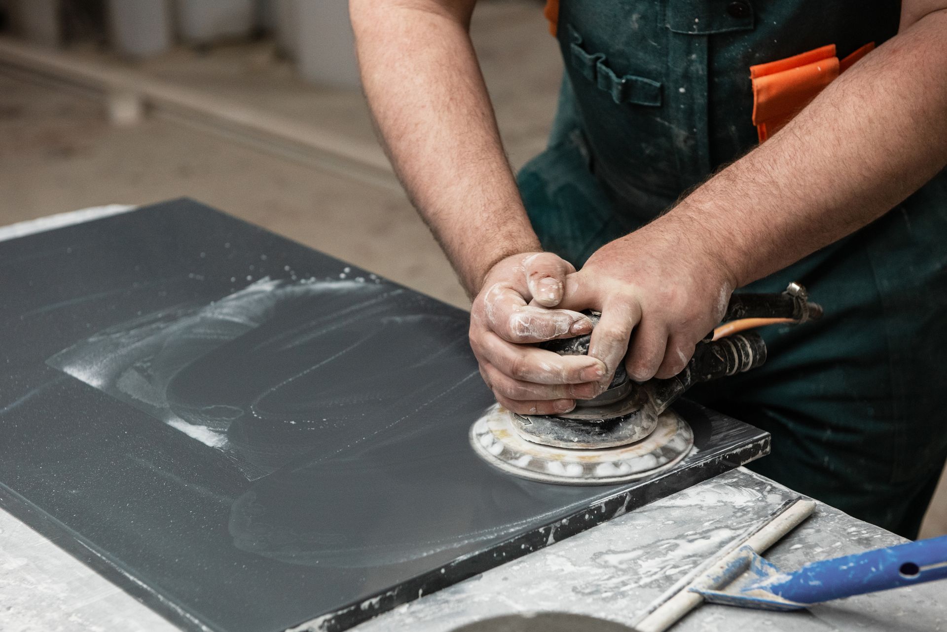 Man sands a dark surface with a power sander, creating dust.