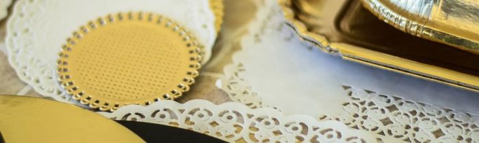 Un primo piano di centrini decorativi di carta bianca sovrapposti a basi rotonde per torte color oro e a un vassoio dorato.
