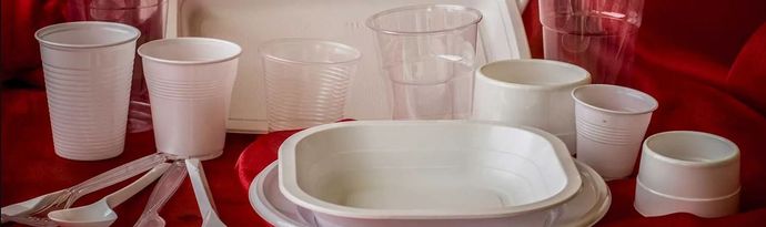 Una collezione di bicchieri, ciotole, piatti e posate di plastica bianca usa e getta, disposti su una superficie rosso scuro.