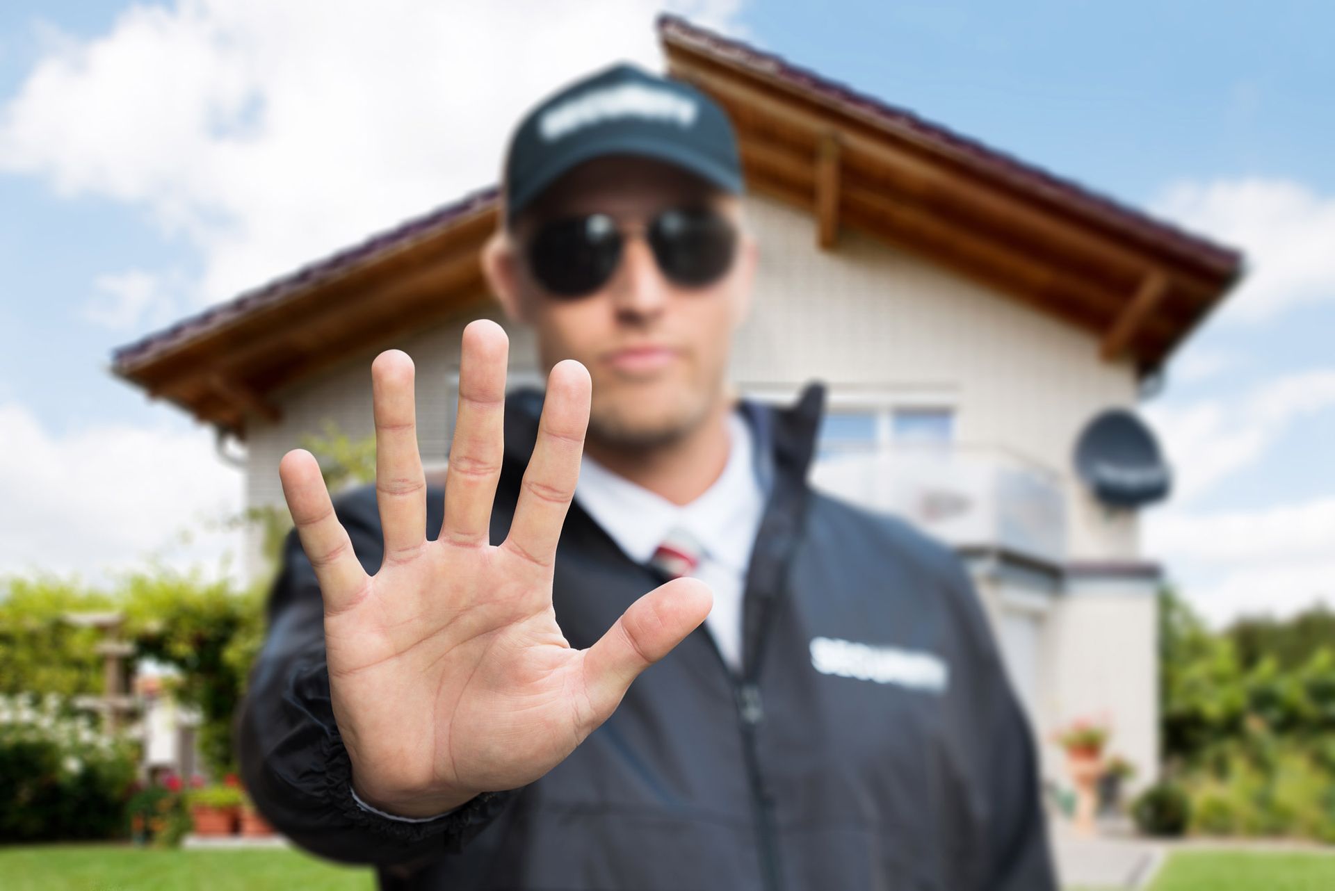 A man wearing sunglasses and a hat is making a stop sign with his hand in front of a house.