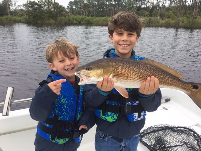 Two boys on a boat proudly display a redfish they caught; both wearing life vests, smiling.