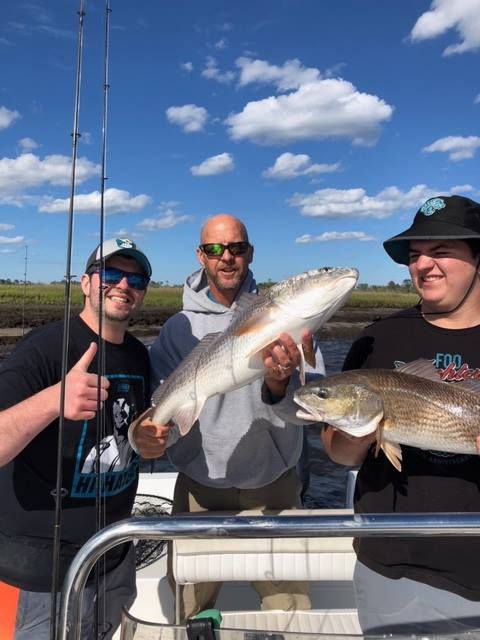 Three people on a boat display their caught fish on a sunny day. Two are holding reddish fish.