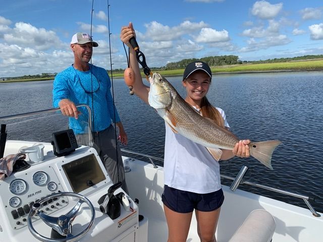 Girl on boat holding up a large redfish caught while fishing, man in background.