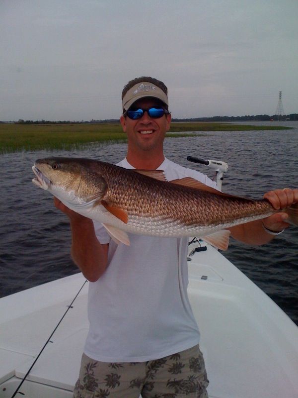 Man holding a redfish on a boat, near marsh grass, smiling.