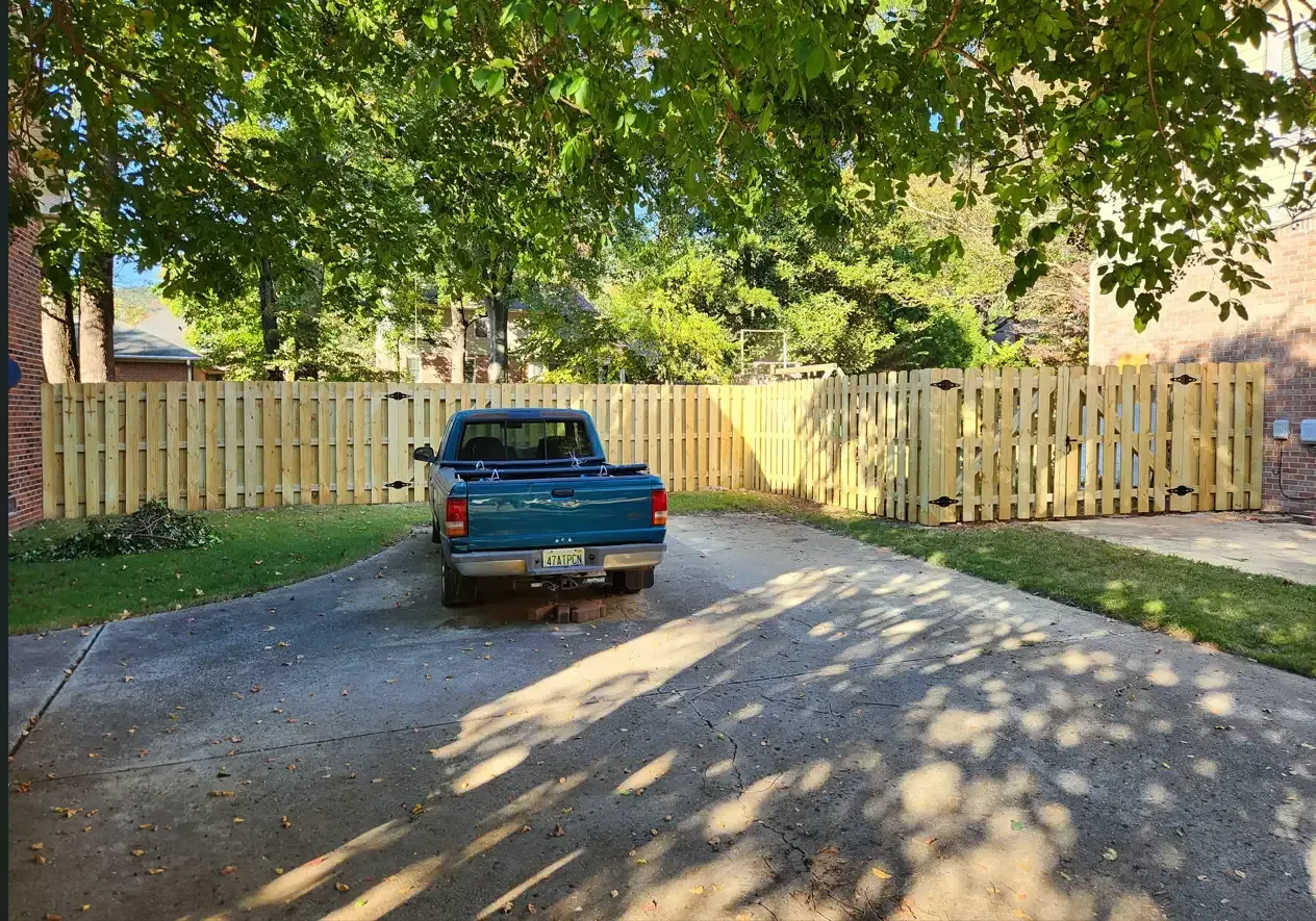 Blue pickup truck parked on a driveway in front of a wooden fence under leafy trees