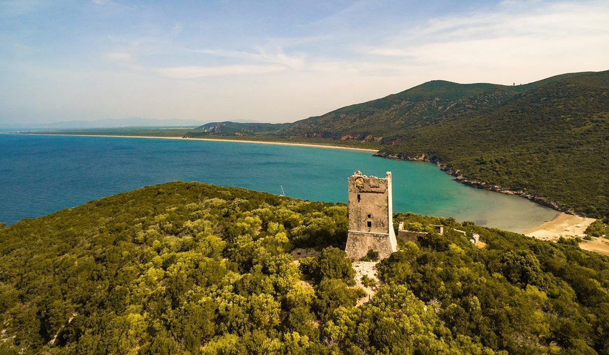 Historic stone watchtower on a green hill above turquoise bay, sandy beach, and distant mountains