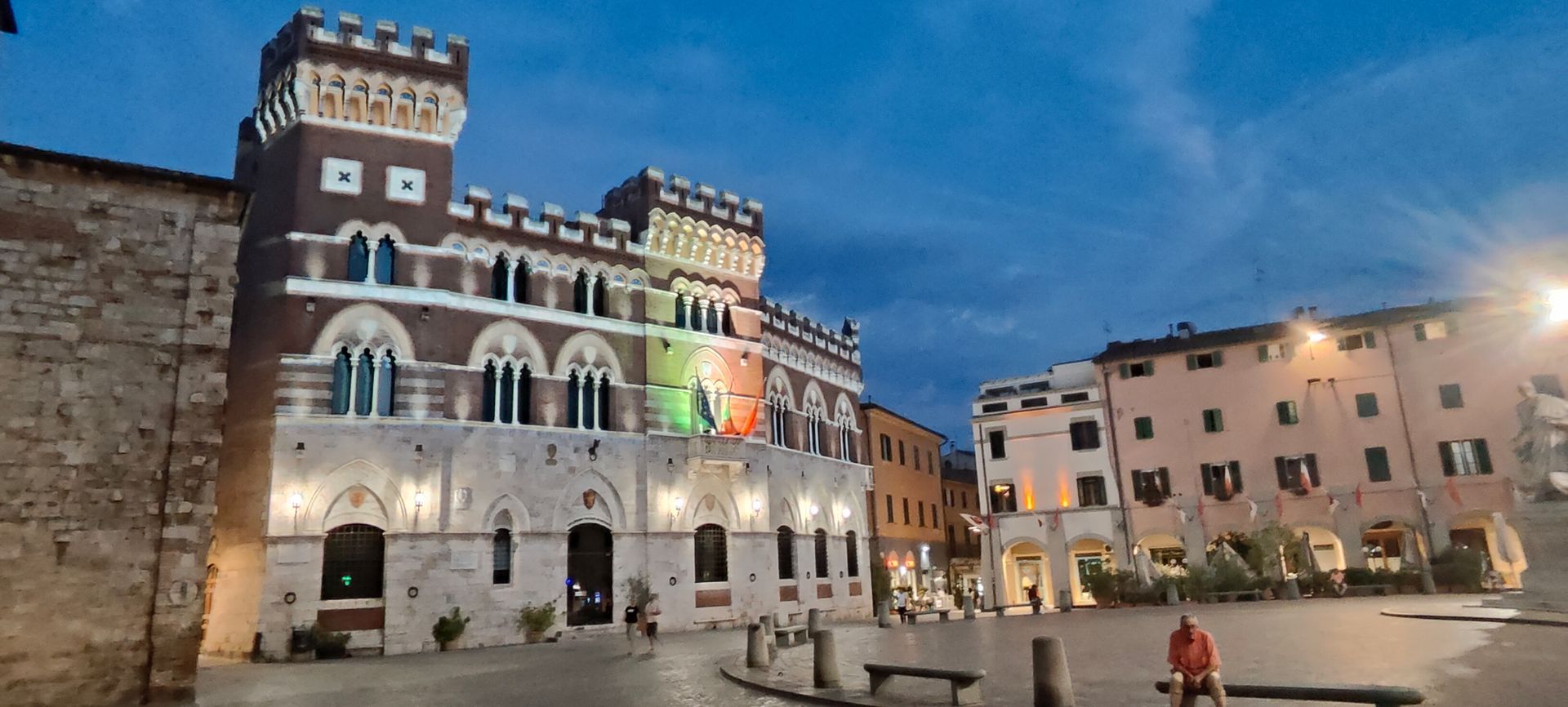 Historic town square in Italy at dusk with lit castle-style building and Italian flag lights