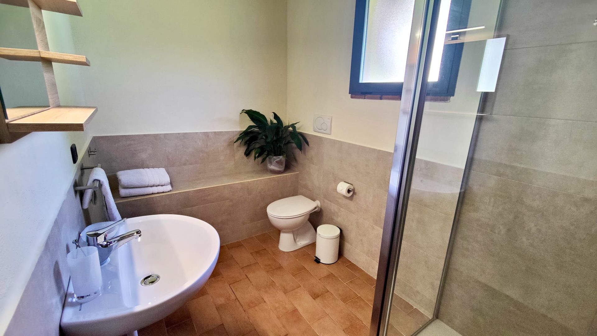 A modern bathroom with beige lower wall tiles and warm-toned floor tiles. On the left, a glass-enclosed shower with chrome fixtures; in the centre, a white sink with a chrome faucet and a glass tumbler on a wall-mounted holder; on the right, a white toilet beside a small trash bin and toilet paper holder. Above the toilet, a shelf holds folded towels and a potted plant, and a frosted window fills the room with soft natural light.