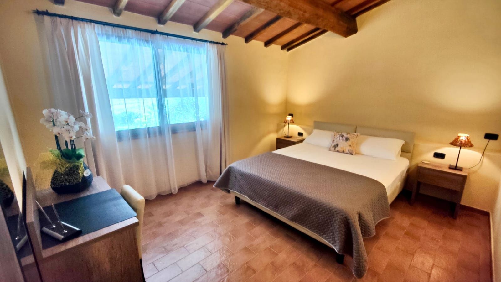A cosy bedroom with a king bed dressed in a grey blanket and two pillows (one with a floral pattern). Exposed wooden beams form the sloped ceiling, and a large window with sheer curtains lets in soft light. Matching bedside tables hold lamps, and a small desk with a chair and a potted orchid sits against one wall. The floor is laid with warm terracotta tiles.