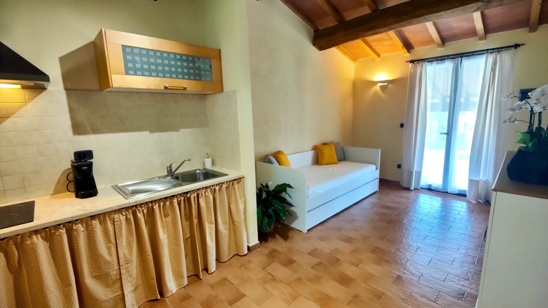 A cosy rustic kitchenette and living nook with exposed wooden ceiling beams and a tiled floor. A countertop holding a sink, stovetop and coffee maker is skirted by a green-and-white chequered curtain beneath frosted-glass cabinets. Beside it, a white daybed with yellow and grey cushions sits under sheer white curtains, with potted plants and a vase of white flowers adding a touch of greenery.