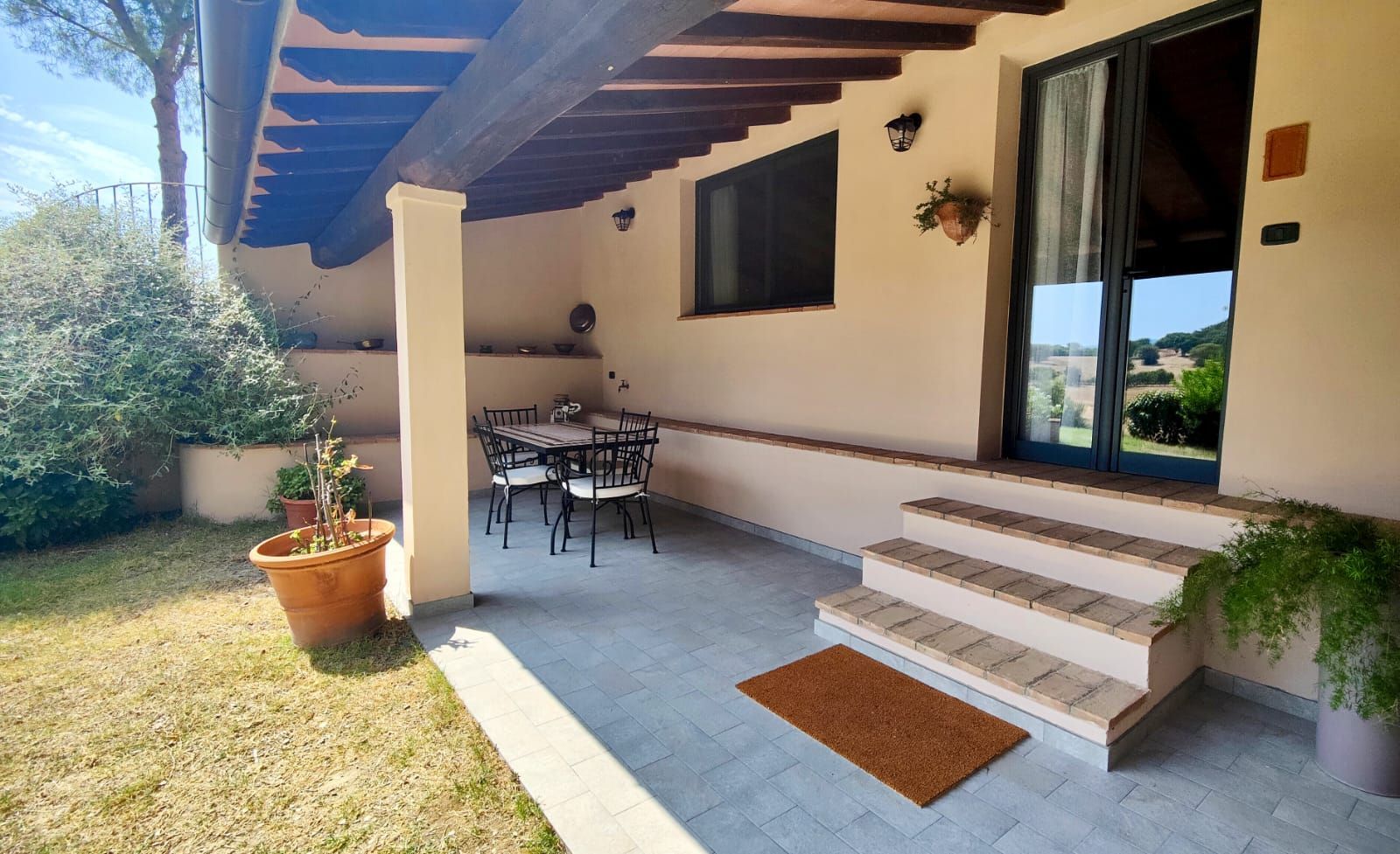 A tiled outdoor patio attached to an apartment, sheltered by a wooden roof on a single column. A small table with four chairs, a potted plant, and a doormat sit in front of large windows and a glass door. Beyond the patio, lush greenery and open fields stretch into the distance.