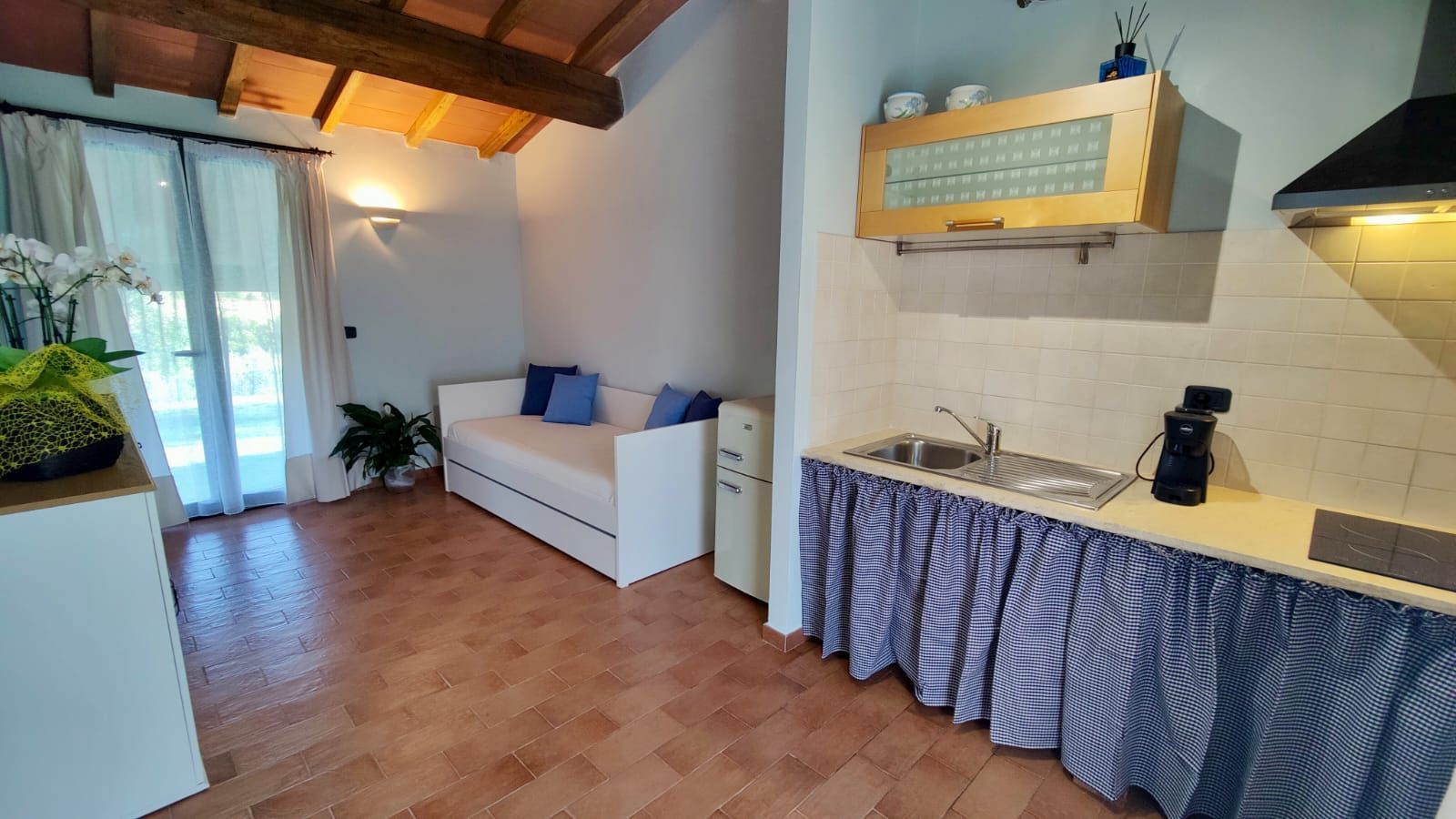 A cosy rustic room with exposed wooden ceiling beams. Along one wall, a small kitchenette features a sink, coffee maker, and stovetop hidden behind a blue-and-white chequered curtain, with glass-fronted wooden cabinets above. Opposite, a white loveseat topped with blue cushions sits beside a compact white refrigerator under a large window dressed in sheer curtains. A small table holding a potted plant and a floral arrangement adds a touch of greenery.