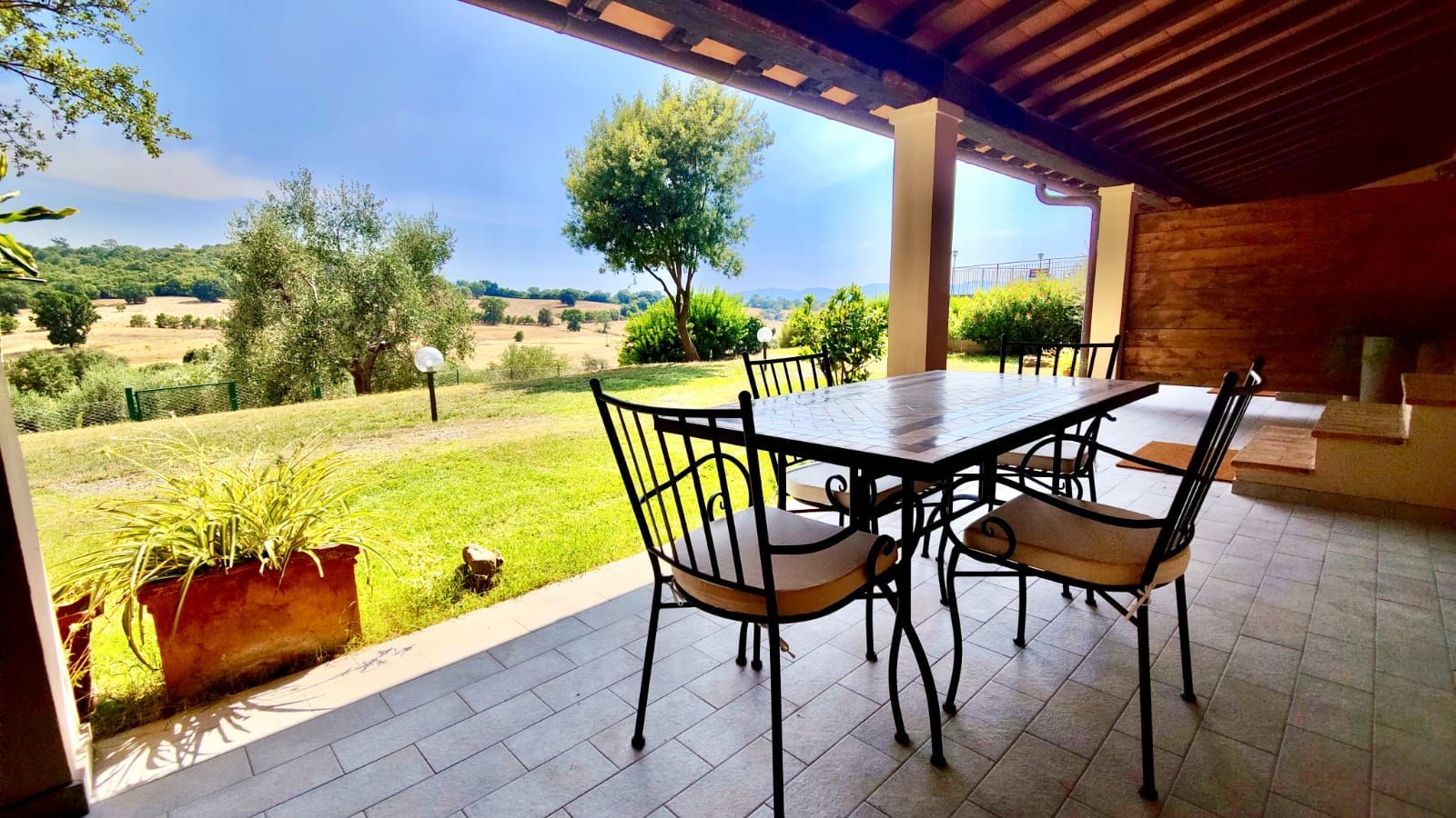 Covered outdoor patio with tiled floor and exposed wooden beam ceiling. A rectangular metal table is surrounded by cushioned chairs. Beyond the patio, a lush green lawn edged with shrubs and trees opens onto rolling hills under a clear, sunlit sky.